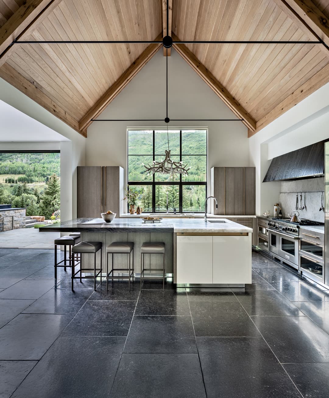 Aspen Colorado mountain luxury kitchen with vaulted timber cathedral ceiling, large island with honed marble countertop, professional Wolf range, oversized window framing aspen valley and mountain view, slate tile floor