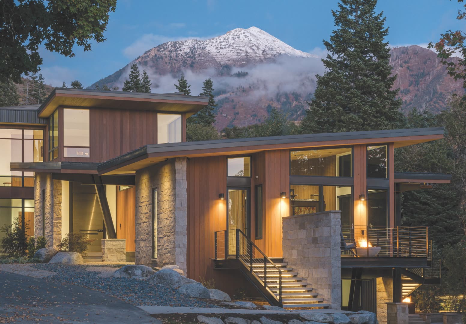 Mountain modern Colorado home exterior at twilight with cedar siding, stacked-stone columns, butterfly rooflines, cable-rail steel staircase, and a snow-dusted Sawatch peak rising behind a clearing of mature spruce and aspen