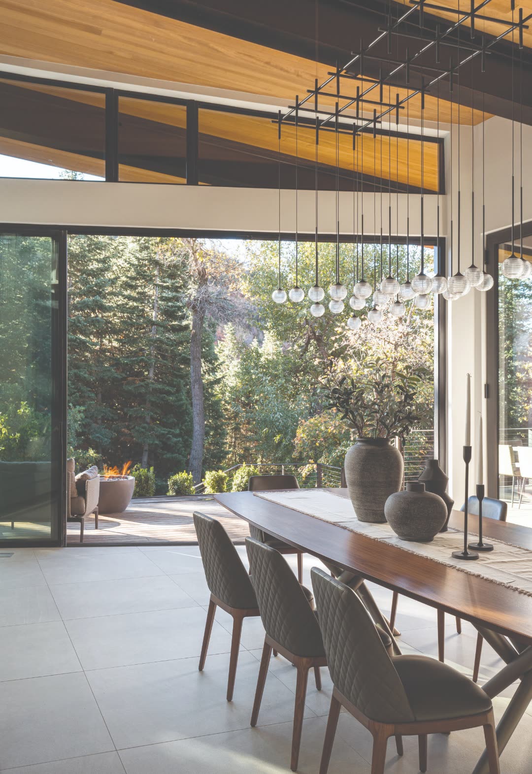 Open-plan Colorado luxury mountain dining room with linear glass-globe pendant chandelier suspended over a long walnut table, leather dining chairs, full-height sliding glass doors opening to a fire pit terrace and forest beyond