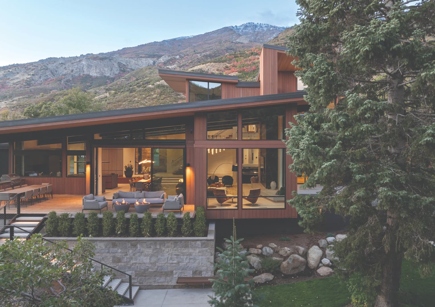 Mountain modern Colorado home exterior at dusk with cedar siding, dramatic cantilevered rooflines, floor-to-ceiling glass walls, stone-clad foundation, and a backdrop of autumn aspens turning red and gold against the foothills