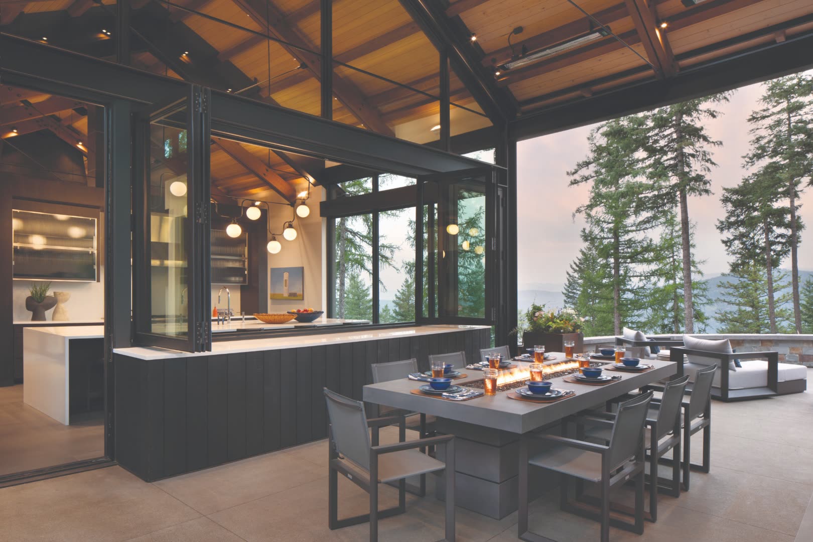 Covered outdoor dining pavilion with retractable accordion-fold glass walls open to a forested Colorado mountain vista at dusk, with a concrete table featuring a built-in fire trough centerpiece and overhead radiant heaters
