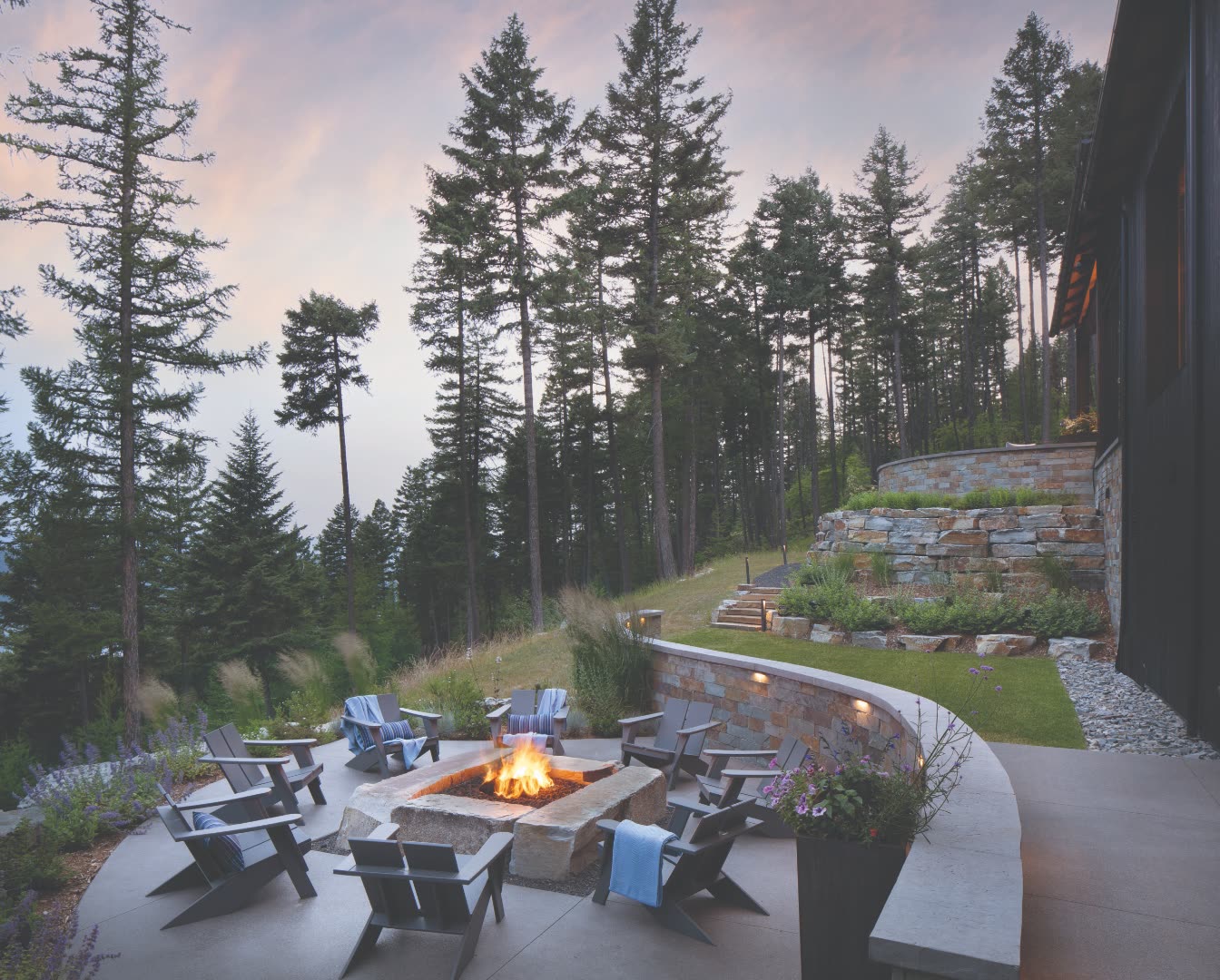 Custom stone-clad outdoor fire pit terrace surrounded by black modern Adirondack chairs, multi-tier dry-stack stone retaining walls planted with native grasses, mature pines, and a soft pink dusk sky
