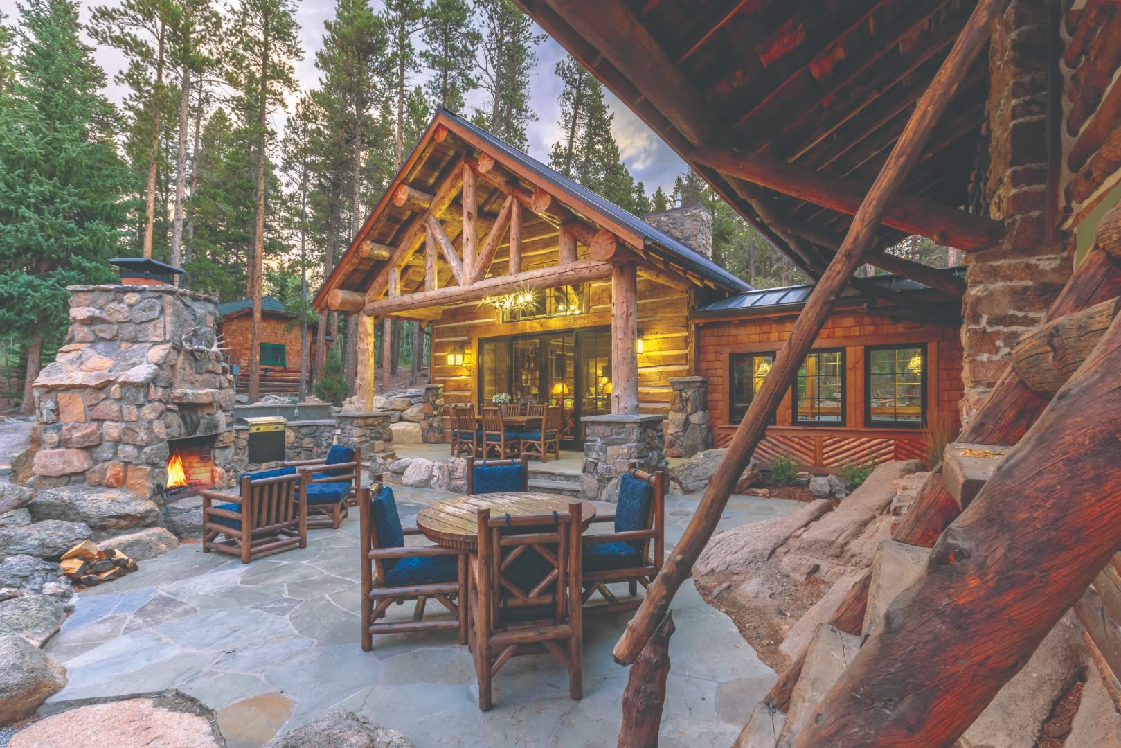 Custom log-and-stone Colorado mountain estate home at dusk with timber-framed gable, large native-stone outdoor fireplace, flagstone patio, hand-hewn log columns, and surrounding stand of lodgepole pine