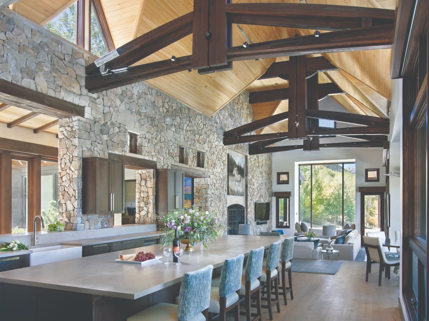 Colorado estate traditional kitchen with full-height hand-laid stone wall, exposed dark-stained timber trusses, cathedral pine ceiling, concrete-topped island with seating for six, and a great room beyond opening to aspens through full-height windows