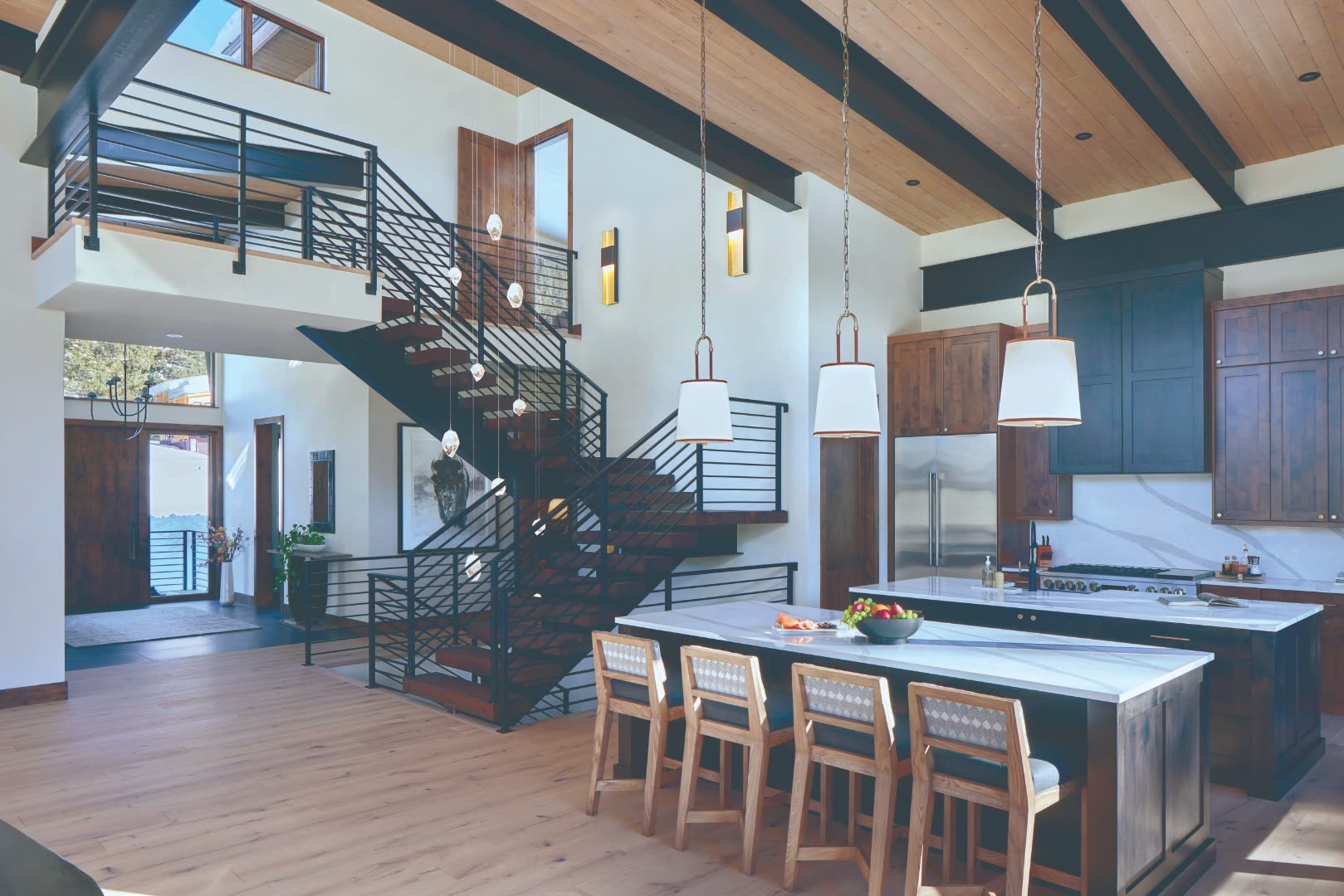 Modern Colorado luxury kitchen with marble waterfall island, walnut cabinetry, exposed steel-and-walnut floating staircase, vaulted timber ceiling, and modern pendant lighting in an open great room