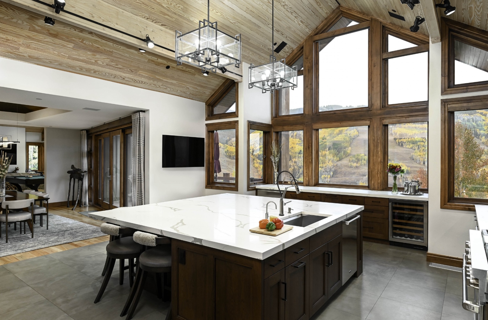 Mountain modern Colorado kitchen with whitewashed tongue-and-groove timber cathedral ceiling, walnut cabinetry, Calacatta marble island with single-sweep waterfall edge, oversized industrial cubic chandeliers, full-height windows framing autumn aspens and ski-run hillside