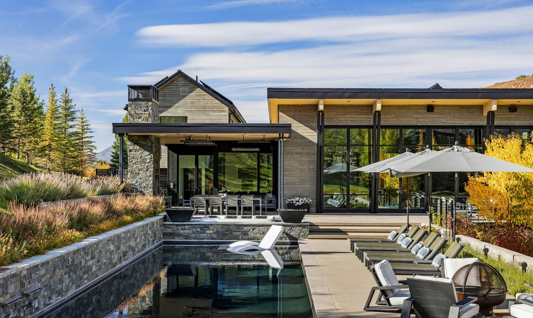 Brothers 59 mountain modern Colorado home exterior with stacked-stone fireplace tower, weathered cedar siding, full-bleed glass great room, infinity-edge pool, and aspens turning gold against the Front Range foothills