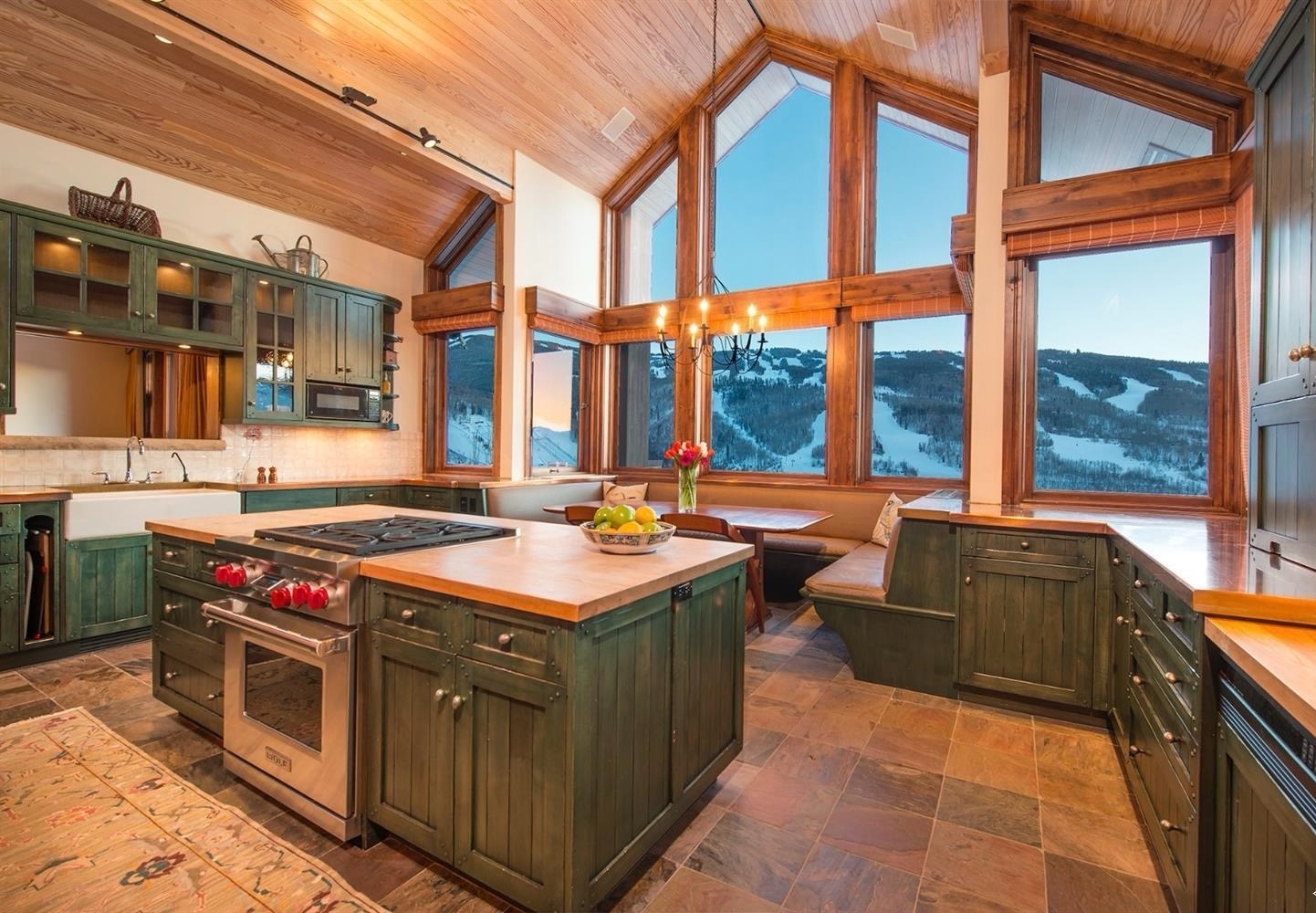 Colorado estate traditional ski-slope kitchen with stained pine cathedral ceiling, green-painted shaker cabinetry, butcher-block island, slate tile floor, banquette nook with antler chandelier, and a wall of cathedral windows framing the Vail ski runs under fresh powder